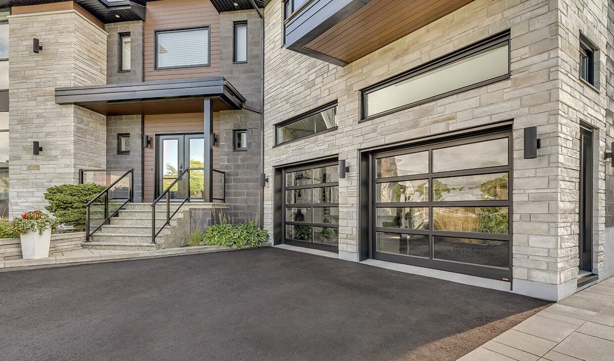 Two full-view garage doors with black aluminium frames and clear glass panels, set into a stone-clad house with a dark asphalt drive.