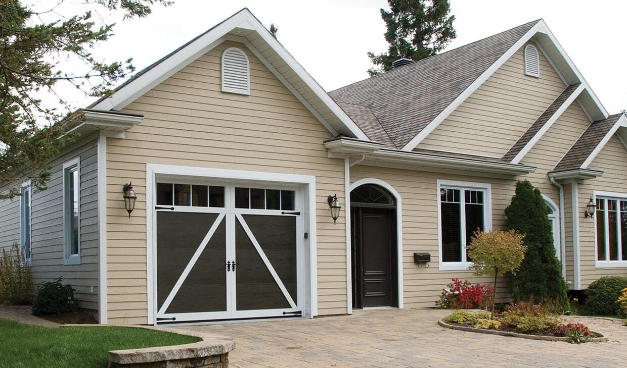 A single dark brown garage door with white crossbuck overlays and strap hinges, with four vertical windows across the top, on a cream sided bungalow.