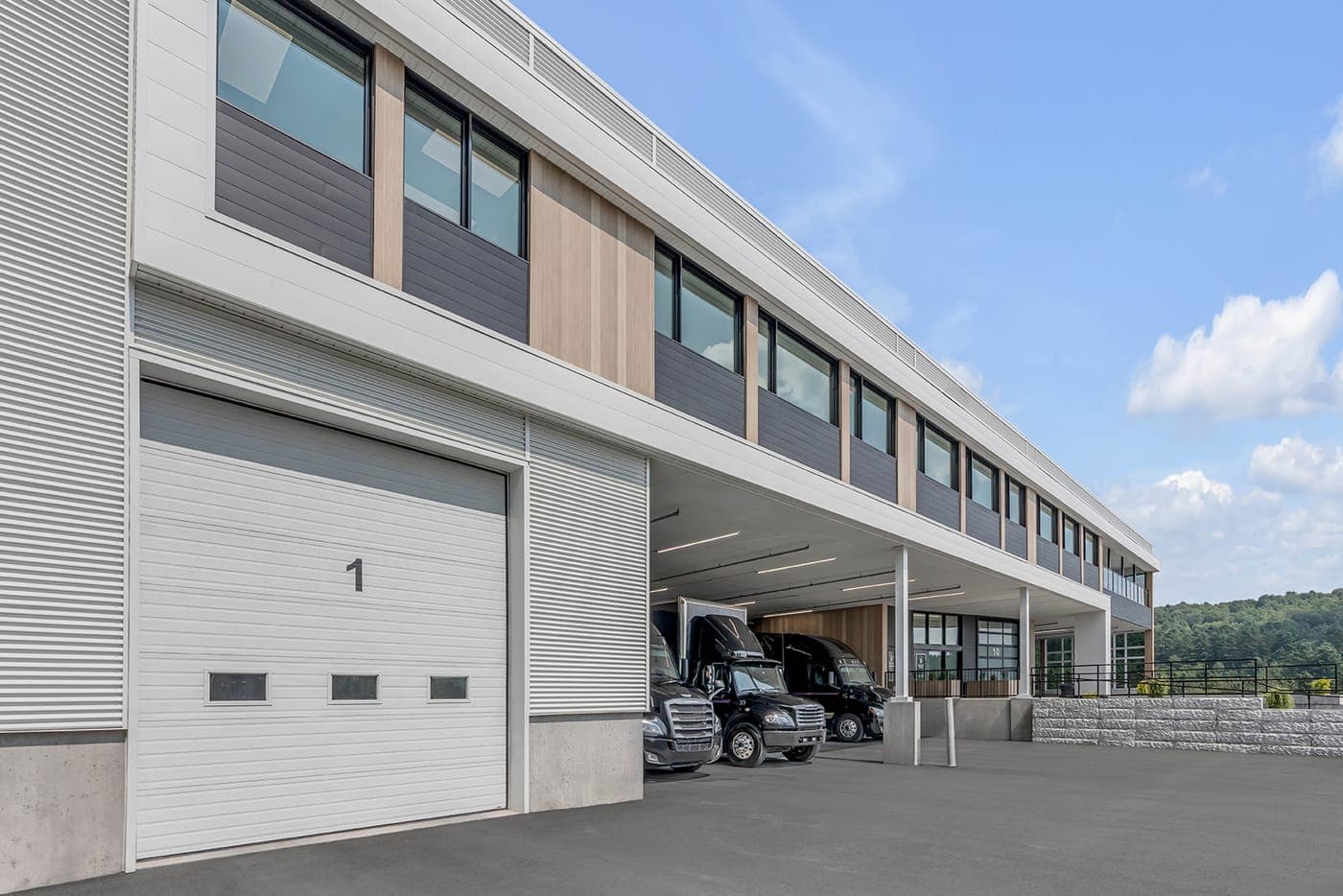 A white insulated sectional door numbered 1 in a ribbed metal commercial building, with transport trucks parked under the canopy beyond it.