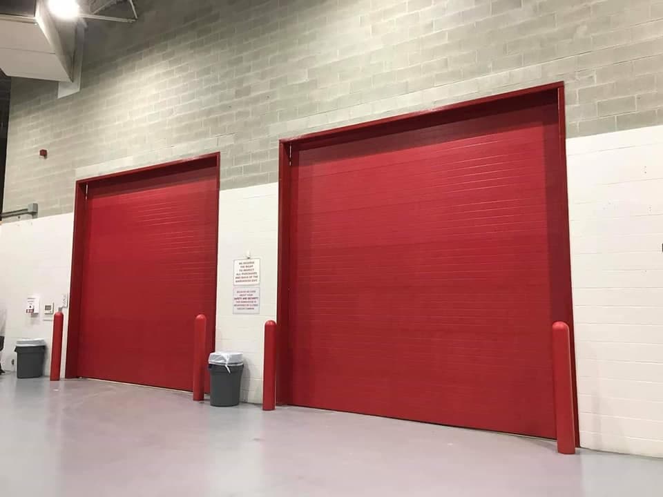 Two red sectional doors on the inside wall of a warehouse, with a red safety bollard at each side of both openings.