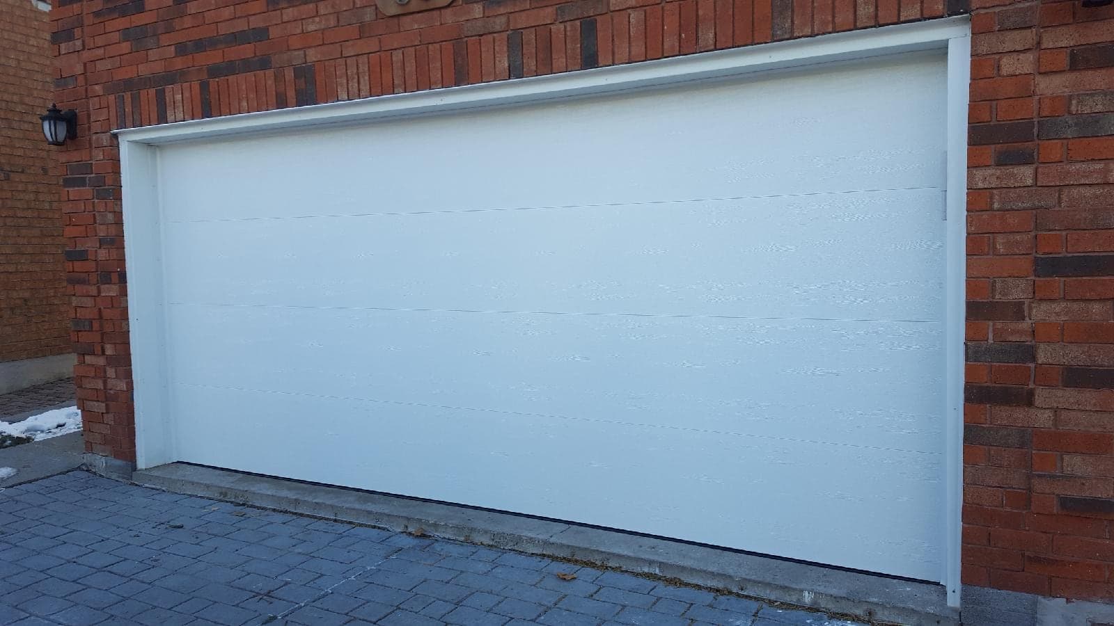 A wide white garage door with a flat face, faint horizontal section lines and no windows, set in a red brick wall above an interlock driveway.