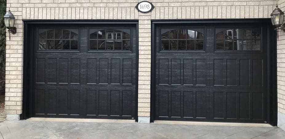 A pair of black woodgrain garage doors with arched divided windows in the top section, on a beige brick house.