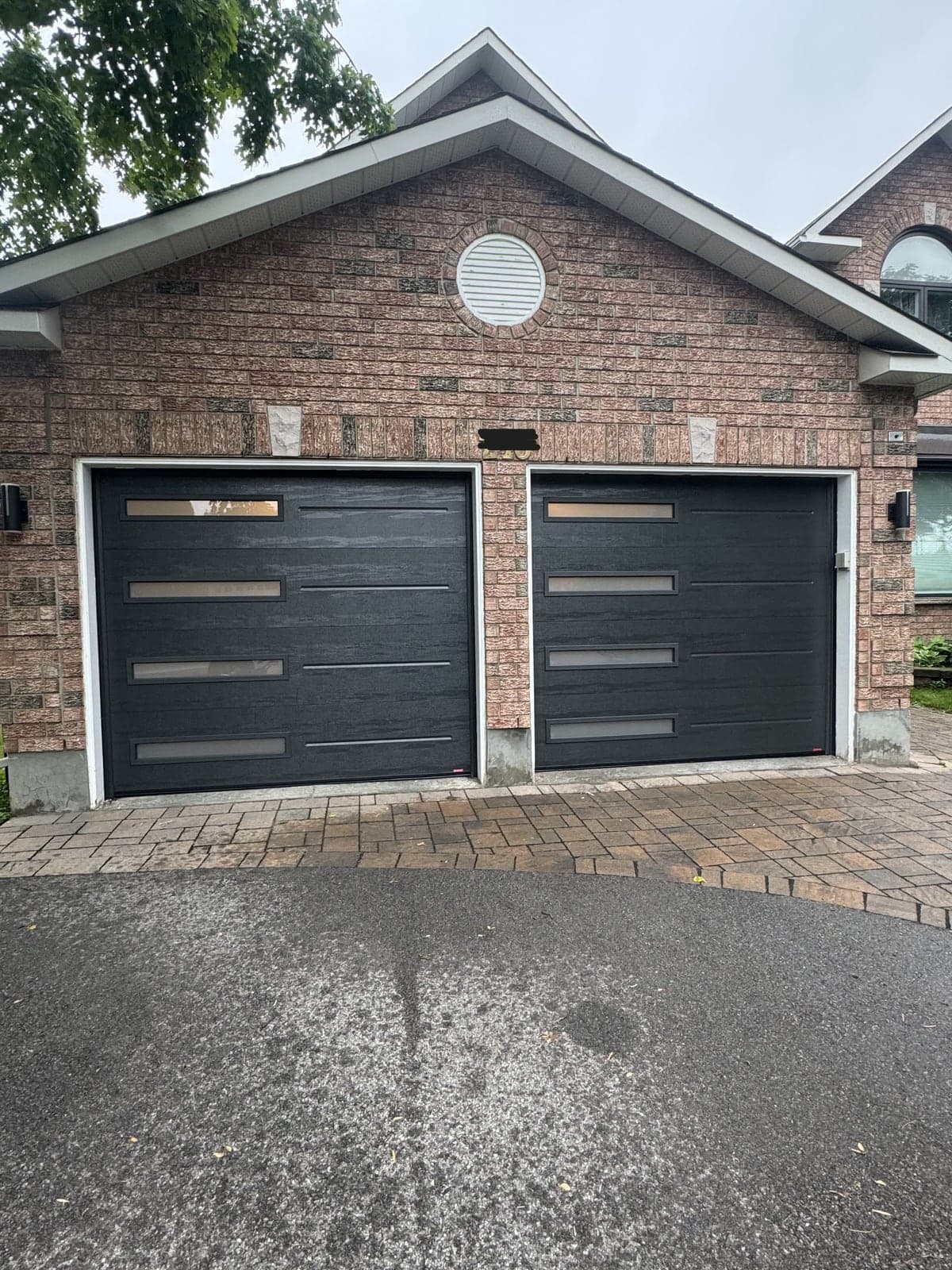 Two black garage doors with horizontal grooves and a column of long narrow windows on each, on a red brick house with a gable above.