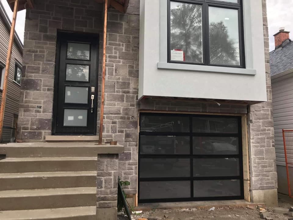 A black full-view garage door with tinted glass panels under a grey stucco bay, beside a black front door with frosted glass inserts.