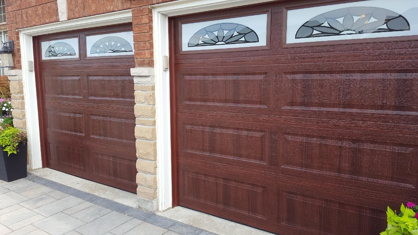 Two brown woodgrain raised-panel garage doors with sunburst-pattern glass inserts across the top, set in brick with a stone pillar between them.