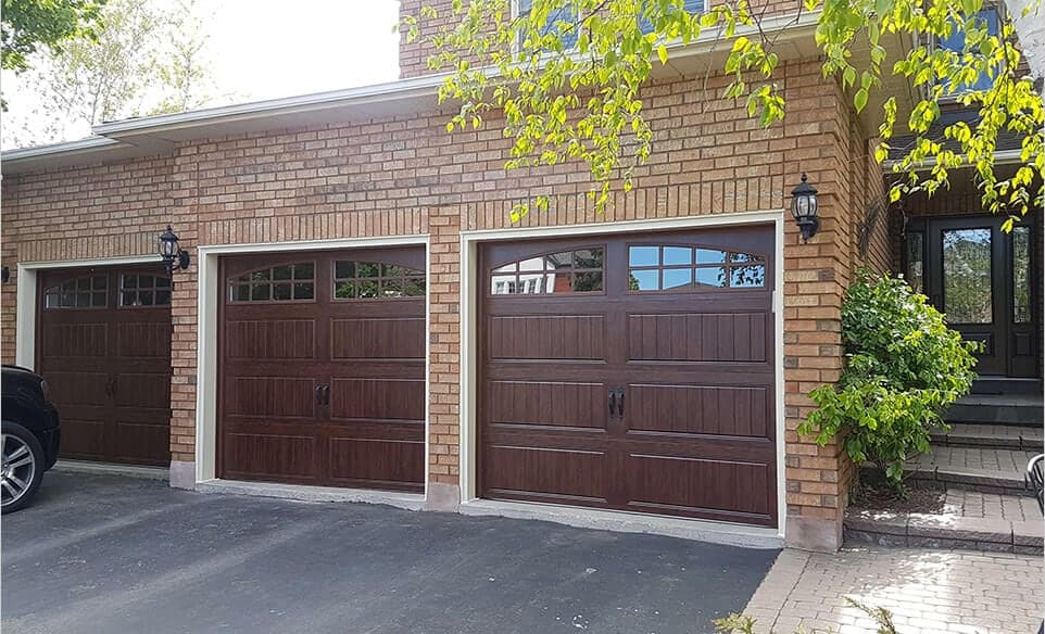 Three woodgrain carriage-style garage doors with arched windows and black handles, on a yellow brick house.
