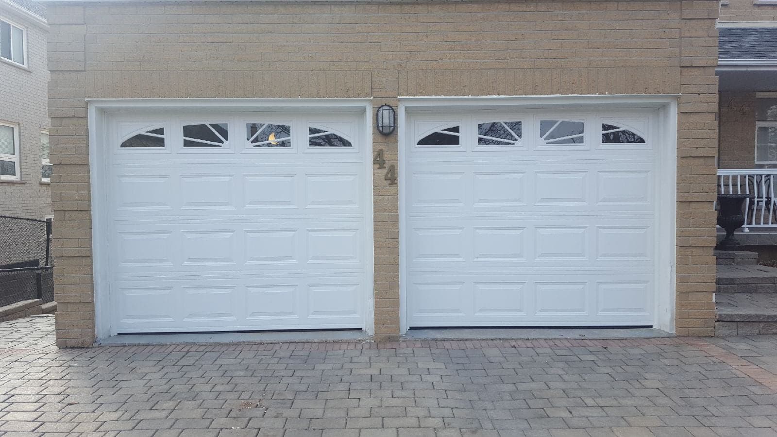 Two white raised-panel garage doors with sunburst-pattern windows across the top, on a buff brick house above an interlock driveway.