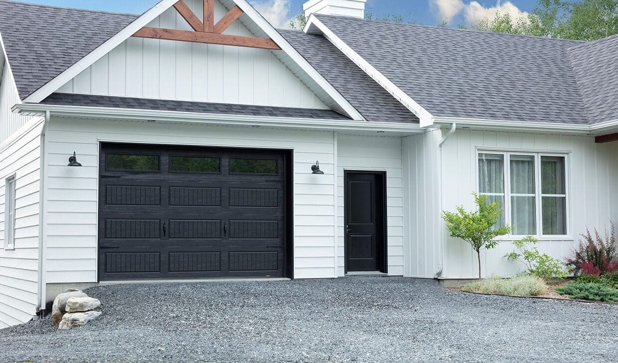A black long-panel garage door with a woodgrain texture and three clear windows in the top section, on a white board-and-batten house.