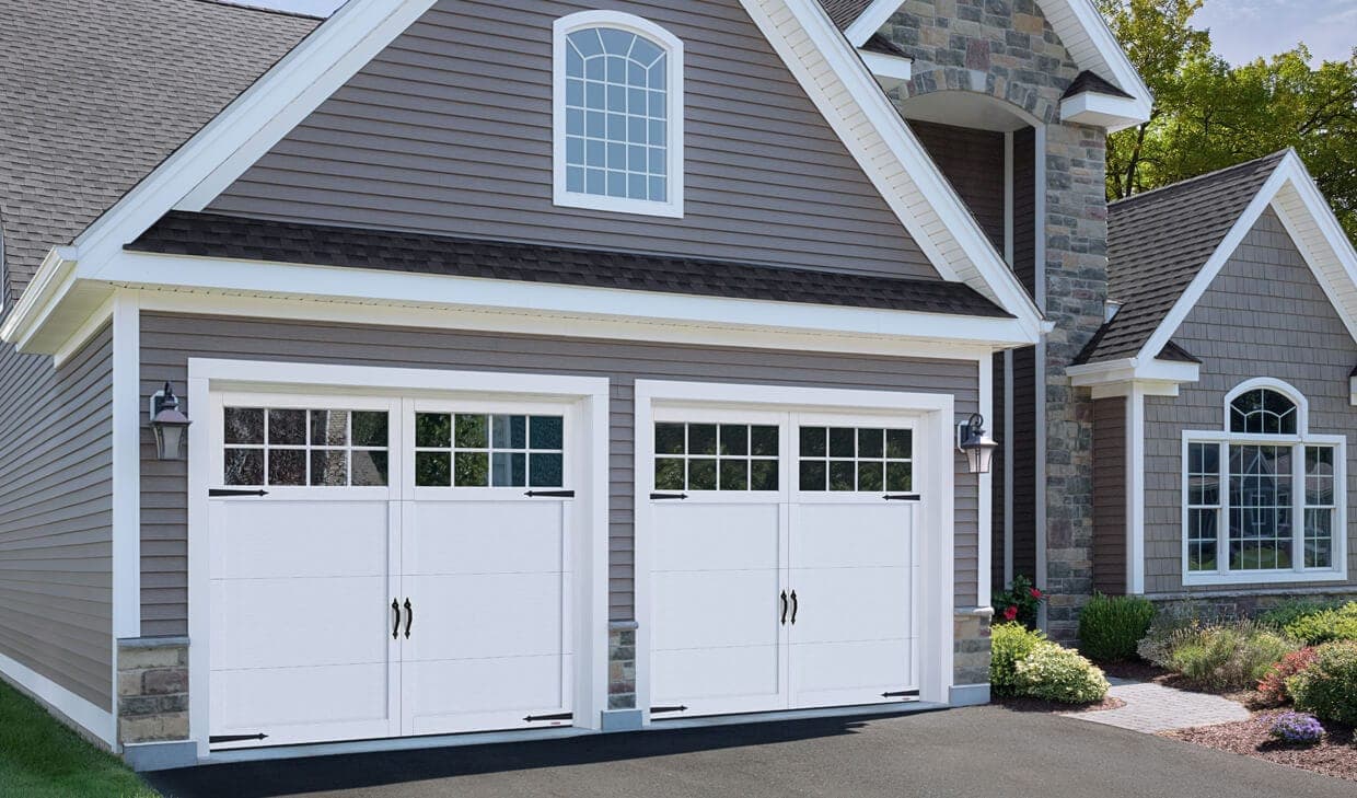 Two white garage doors with applied overlays, black strap hinges and handles, and a row of divided windows in the top section, on a shingled suburban house.