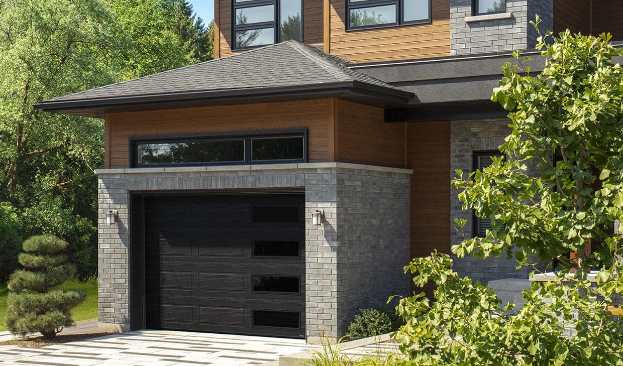 A black garage door with wide horizontal sections and a column of small dark windows set to the right, in a grey brick opening under a timber soffit.