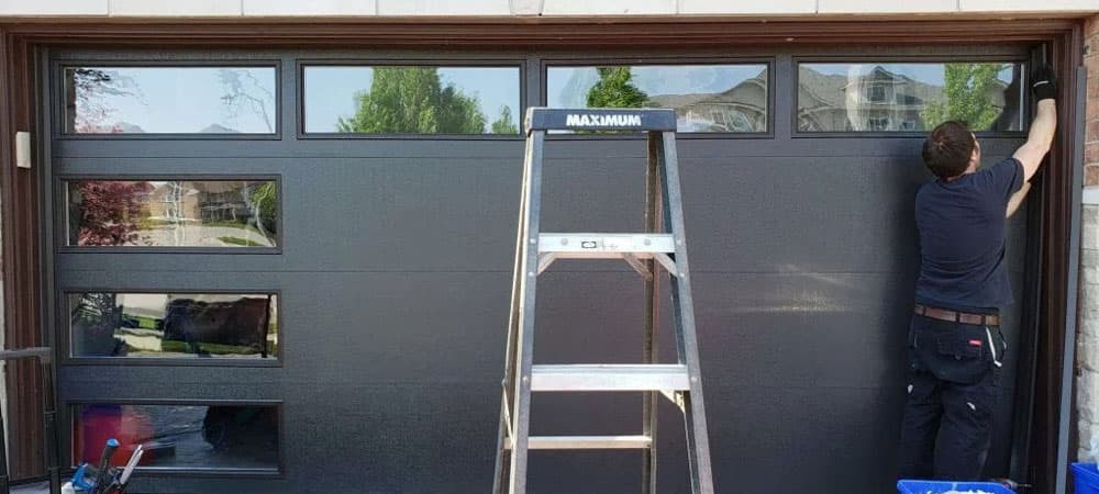 A technician working at the edge of a dark grey sectional garage door with a row of windows in the top panel.