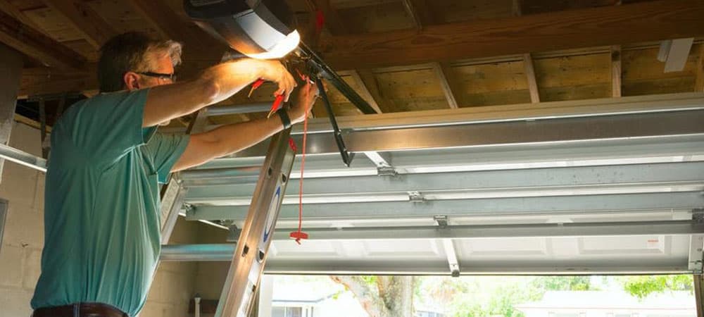 A technician on a stepladder working on a garage door opener mounted to the ceiling joists.