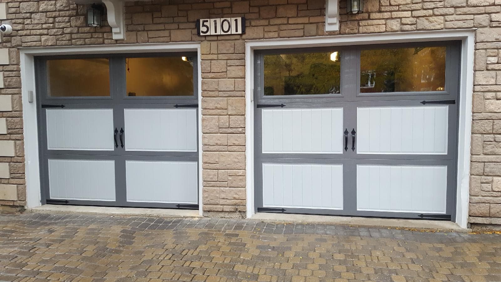 Two carriage-style garage doors with grey frames, pale grey panels, black handles and strap hinges, and clear windows across the top, in a stone wall.