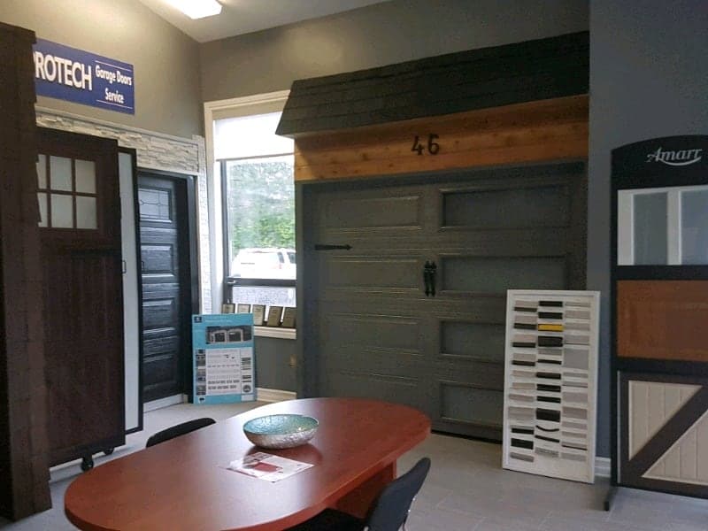 A charcoal long-panel garage door with black handles and a strap hinge, built into a display wall in the Protech showroom, with a colour sample board and a meeting table in front of it.