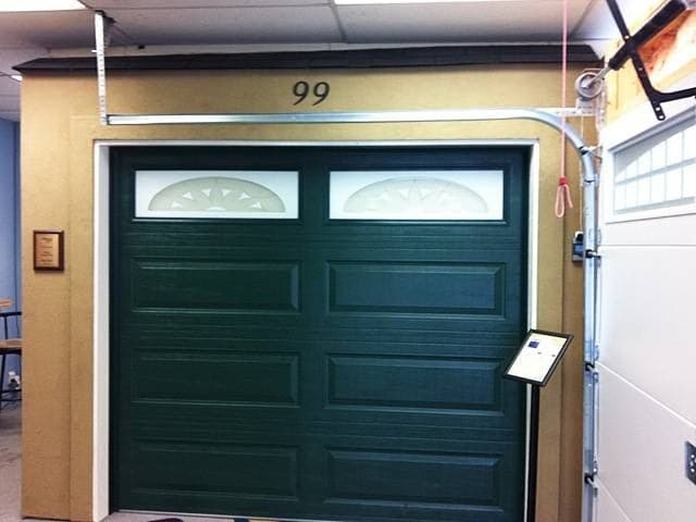 A dark green raised-panel garage door with two sunburst-pattern glass inserts, mounted on a display wall in the showroom with its track and an information stand beside it.