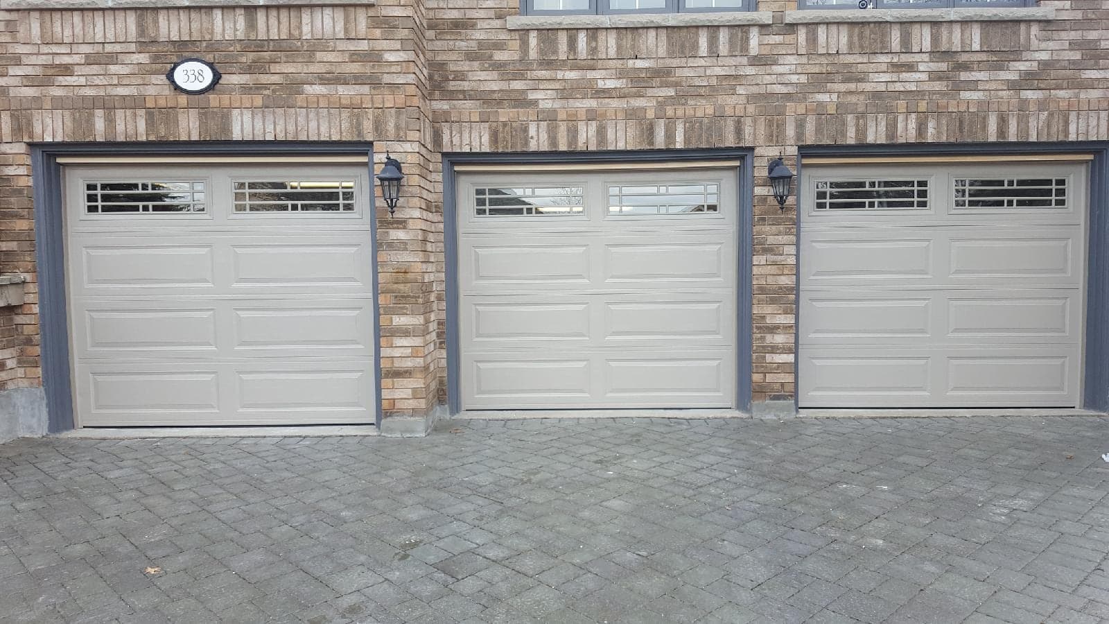 Three beige long-panel garage doors with a row of divided windows along the top, set in a brick wall with grey trim above an interlock driveway.