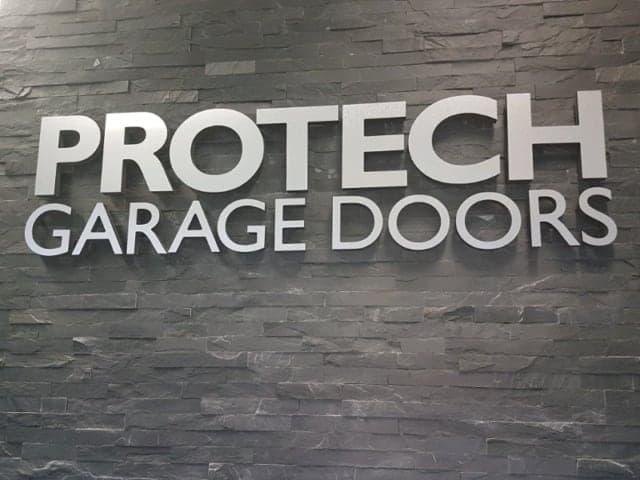Brushed metal letters reading Protech Garage Doors, mounted on a dark stacked-stone wall.