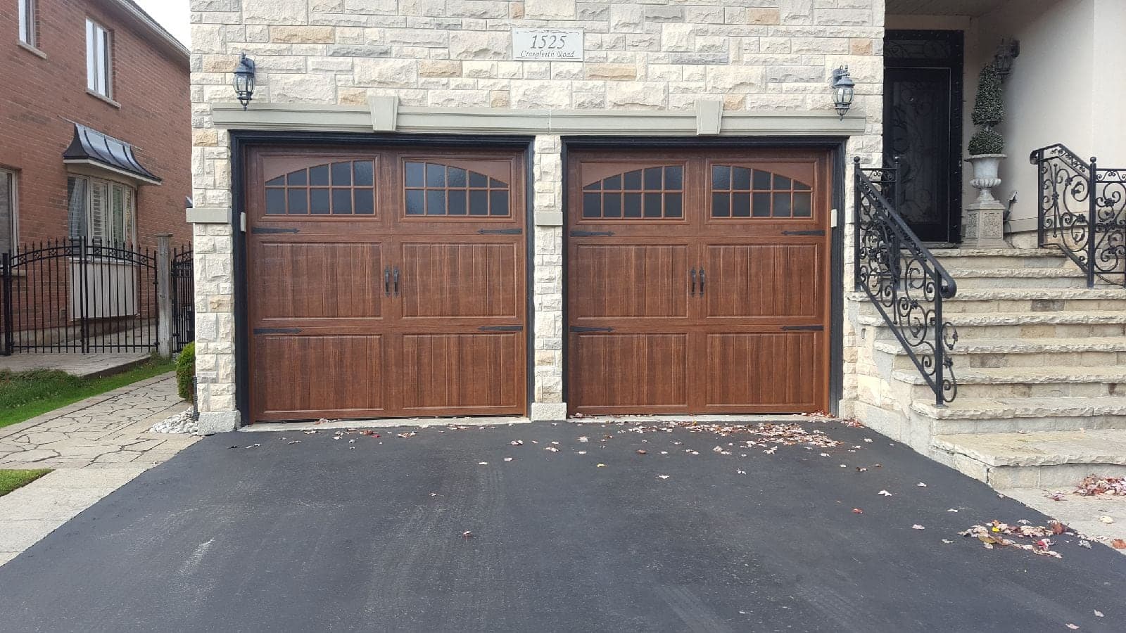 Two woodgrain carriage-style garage doors with arched divided windows, handles and strap hinges, in a pale stone wall beside a front staircase with iron railings.